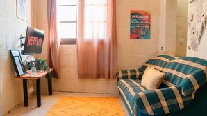 Un espacio de sala cómodo con un sofá de tapiz azul y verde, una pequeña mesa de madera con un marco y una planta, una alfombra anaranjada grande y un televisor montado en la pared que muestra el logo de Netflix.
