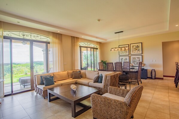 Amplia sala de estar con un sofá grande, una mesa de centro y sillones de mimbre. Hay un comedor con una mesa y sillas. La habitación tiene ventanas grandes con cortinas, que ofrecen vistas de la vegetación exterior.