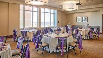 A banquet hall with round tables, chairs, and purple sashes, featuring large windows and ceiling lights.