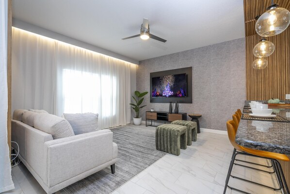 A modern living room with a sofa, ottoman, dining table, and chairs. There is a ceiling fan, a flat-screen TV mounted on the wall, and a potted plant. - 3119- Great Family Home near Disney, private pool, Storey Lake Resort. (Kissimmee)