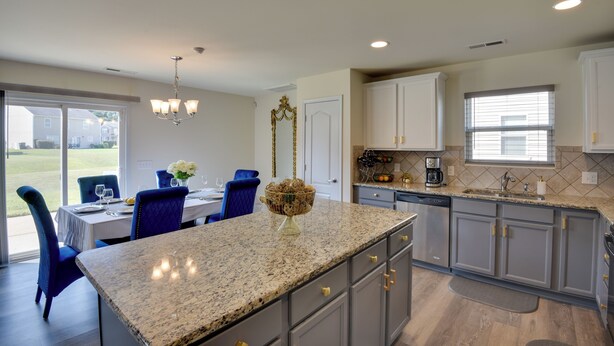 A modern kitchen with granite countertops, built-in cabinets, and a dining area with blue chairs and a table set for four. - Spacious Gastonia Home w/ Pool Access! (Gastonia)