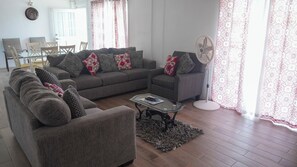 Un salon avec un canapé, un fauteuil et une table basse. On y trouve également un ventilateur et un tapis sur le sol.