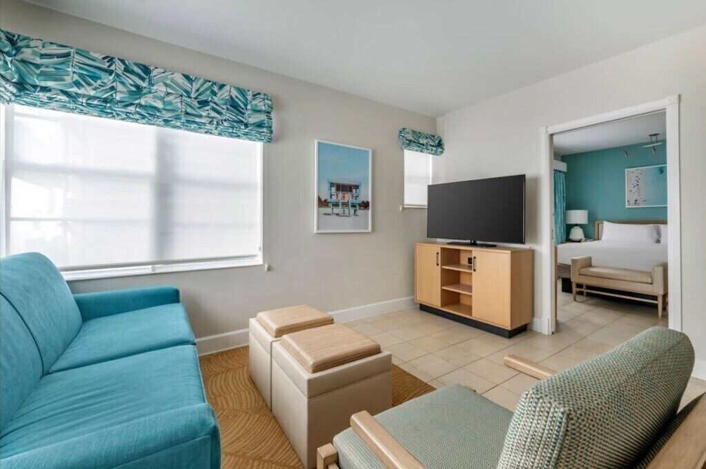 A modern living room with a sofa, ottomans, a TV on a wooden stand, and a bed in the adjacent room.