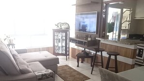 Una sala de estar con sofá, televisión, ventilador de techo y una zona de cocina con mostrador y taburetes.