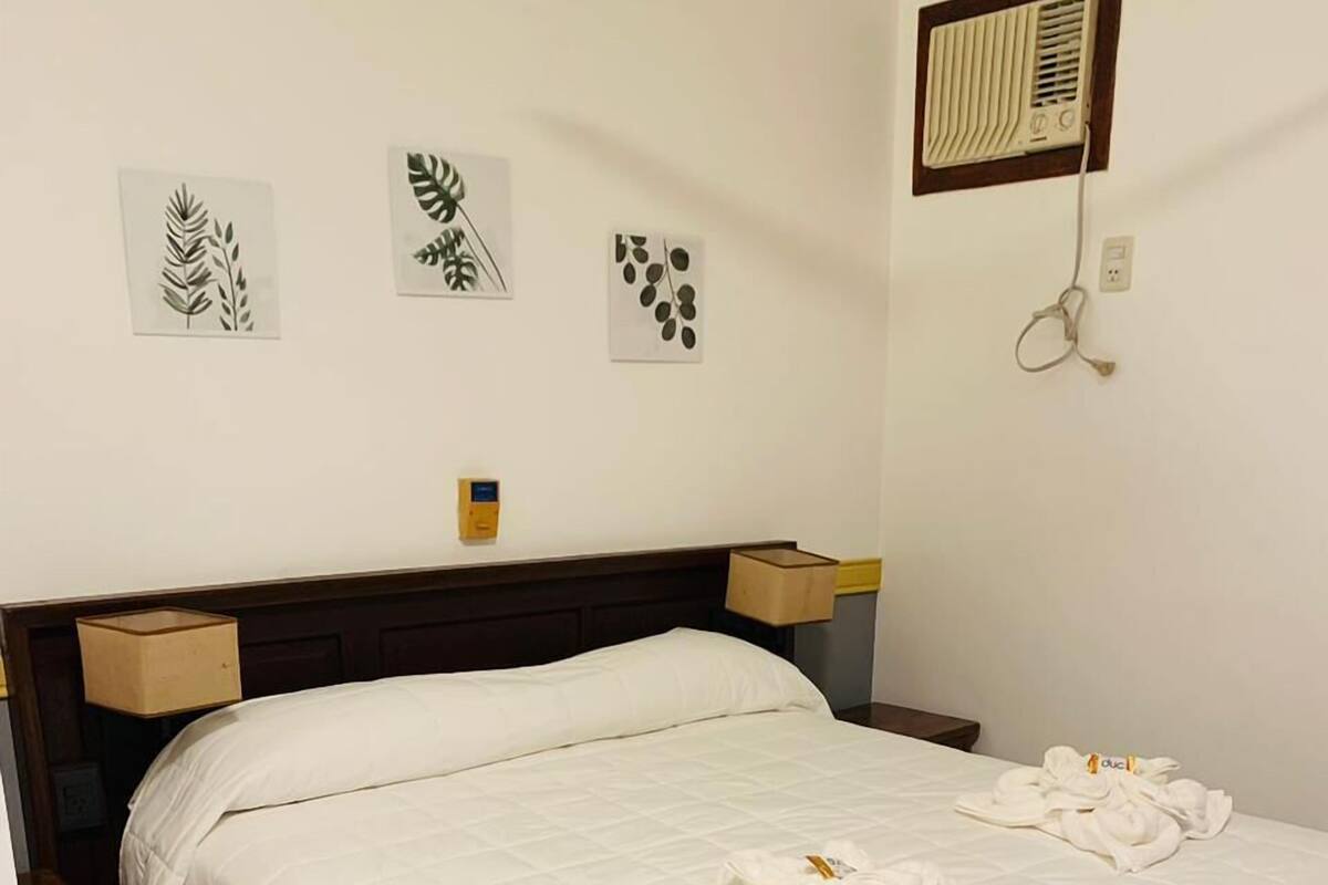 A neatly made bed with white bedding and towels, a wooden headboard, bedside tables, and a wall-mounted air vent.