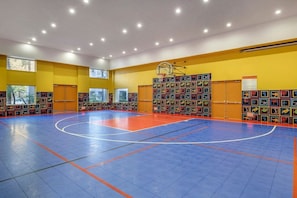 A basketball court with a blue and orange floor, surrounded by yellow walls and windows.