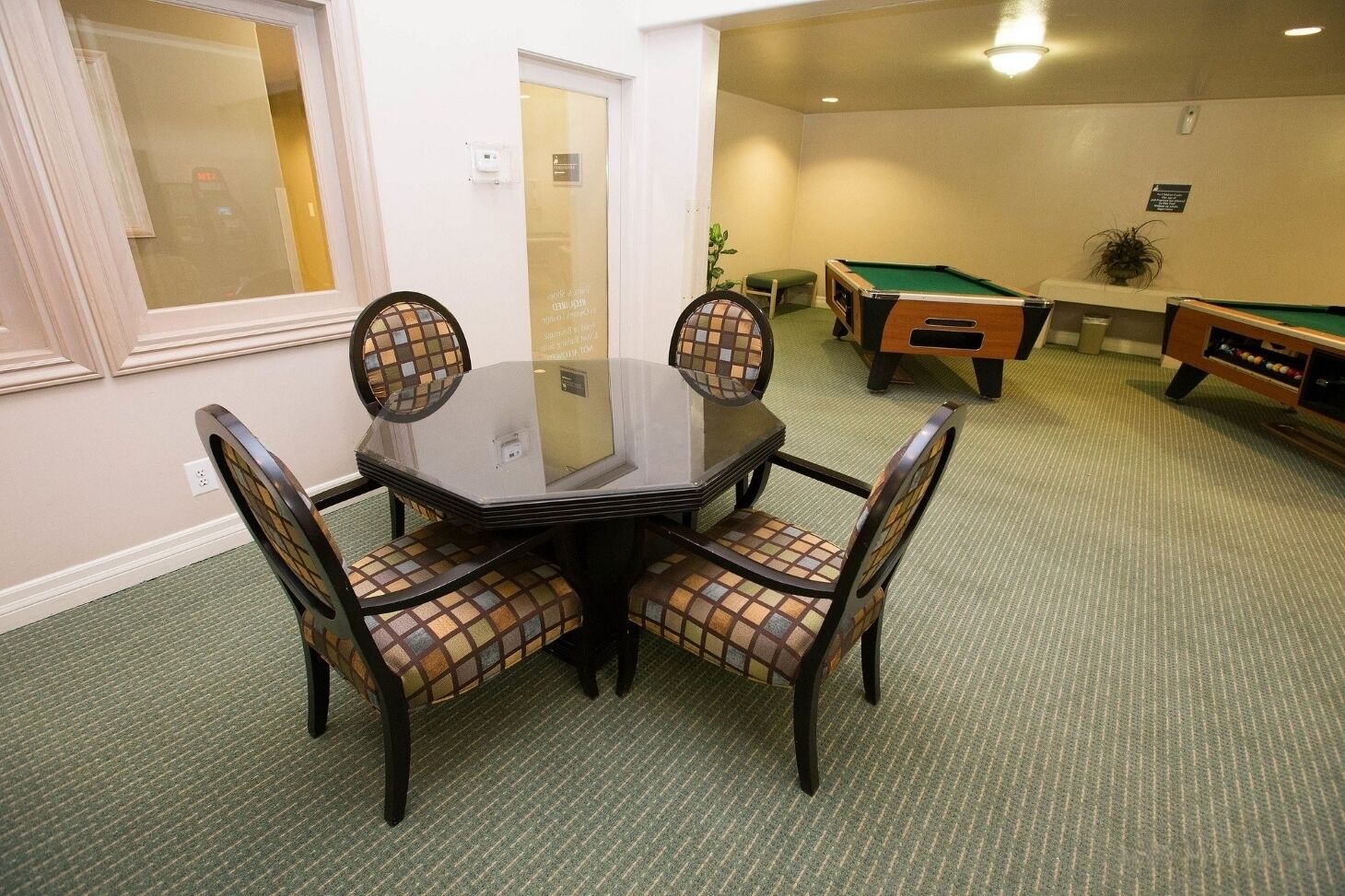 A room with a glass-top table surrounded by four chairs with checkered upholstery, a pool table, and a green carpet.