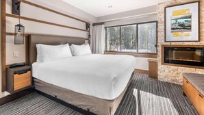 Une chambre d’hôtel moderne dotée d’un grand lit, d’une cheminée intégrée, d’une grande fenêtre donnant sur la forêt et d’un mur décoratif en pierre.