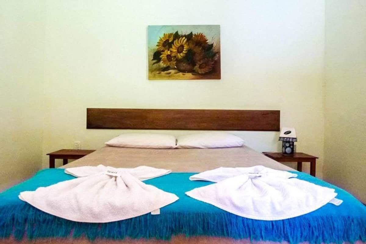 A bed with a blue and pink bedspread, two towels on the bed, a wooden headboard, a nightstand with a lamp, and a painting on the wall.