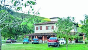 A two-story building with a red-tiled roof and a small porch area. There are several cars parked in front, and a grassy area with trees and shrubs. - Pousada Rancho da Mata (Valenca)