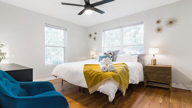 A bedroom with a bed, two bedside tables, a blue chair, and a ceiling fan. - Modern Cottage by Hospital, King Bed / Glass Shower (Joplin)