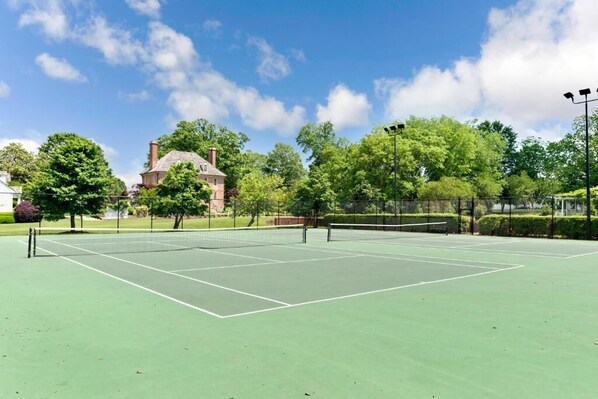 An outdoor tennis court with a net, surrounded by trees and a building in the background.