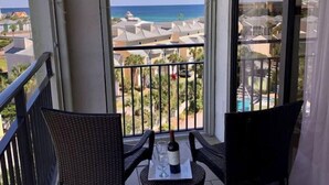 Un balcon avec des fauteuils en osier, une table avec une bouteille et des verres, et une vue sur l’océan et les bâtiments du complexe hôtelier.