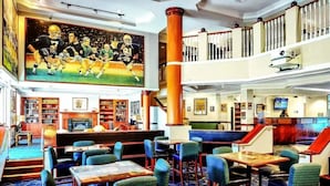 Un vestíbulo de hotel con un gran mural con temática deportiva, varias zonas de estar, una chimenea y una escalera a un nivel superior.