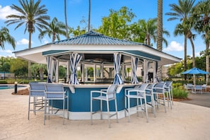 Eine überdachte Bar im Freien mit Sitzgelegenheiten, umgeben von Palmen und einem Pool.