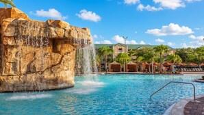 A pool with a waterfall feature, surrounded by lounge areas and a building with a green roof.