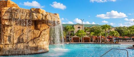 A pool with a waterfall feature, surrounded by lounge areas and a building with a green roof.