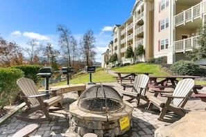 A patio area with Adirondack chairs, a stone fire pit, picnic tables, and a multi-story building with balconies.