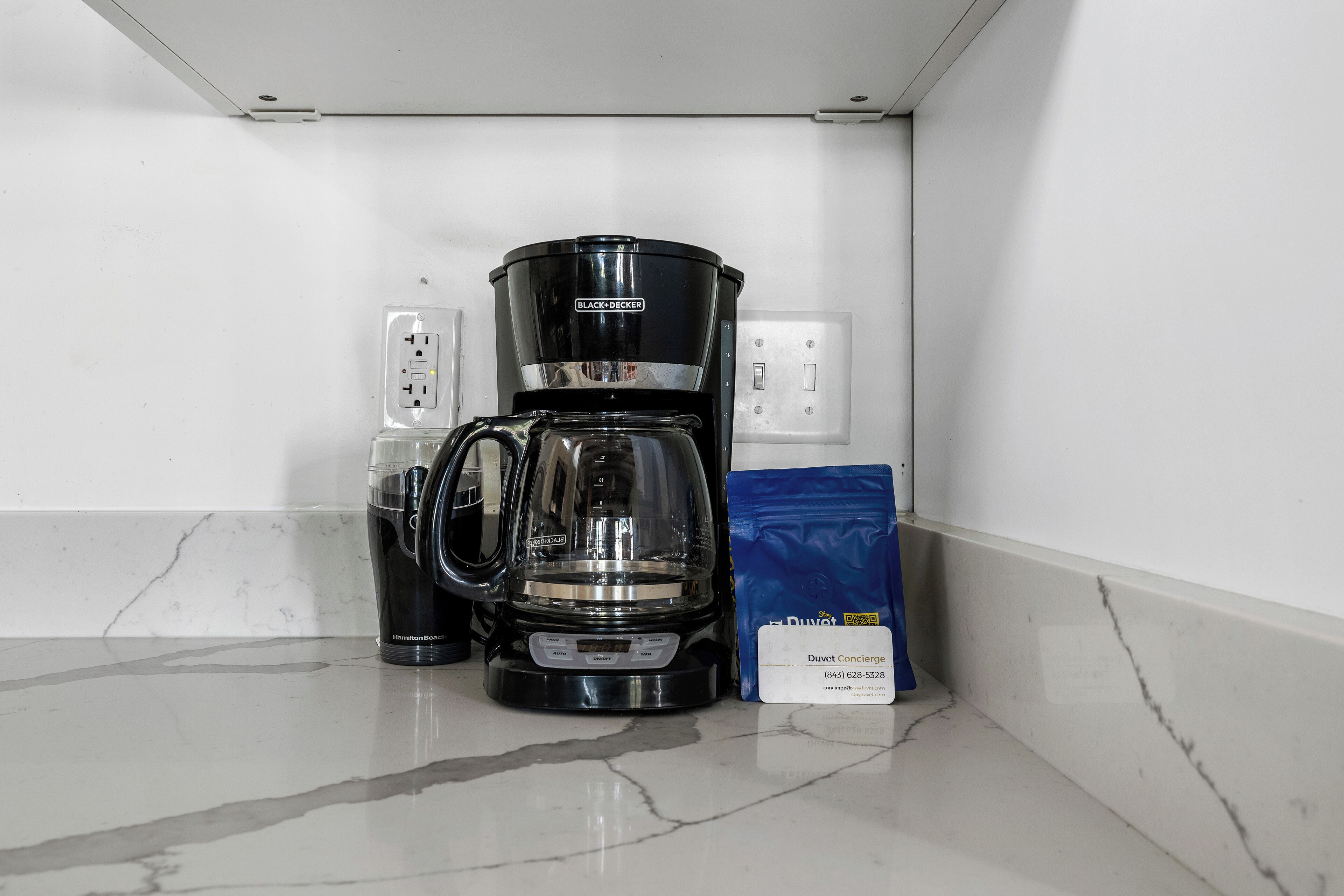 A black coffee maker on a marble countertop next to a blue bag of coffee and a glass carafe.
