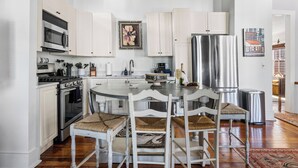 Una cocina con armarios blancos, electrodomésticos de acero inoxidable, una mesa redonda con sillas blancas y suelo de madera.