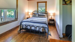 A bedroom with a metal bed frame, wooden flooring, a window with blinds, and a nightstand with a lamp. - Creek, Trails & Falls! Rural Red River Gorge Cabin (Campton)