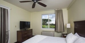 A hotel room with a bed, a television, a window with curtains, a ceiling fan, and a nightstand with a lamp.