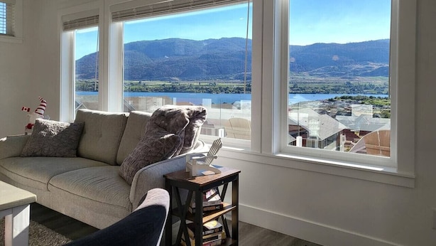 A living room with a sofa and armchairs, large windows with a view of mountains and a lake, and a small table with a lamp.