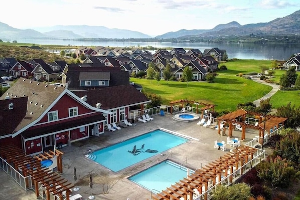 A residential area with a large swimming pool, multiple houses, and a lake in the background. - Eagle View Cottage #243 (Osoyoos)