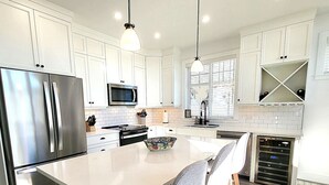 A modern kitchen with white cabinetry, stainless steel appliances, a central island, pendant lighting, and a window with blinds.