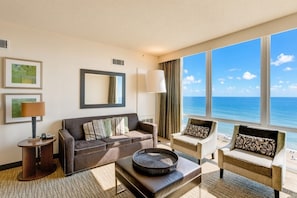 A hotel room with a large window offering a view of the ocean, a sofa, a chair, a lamp, and a small table with a tray.