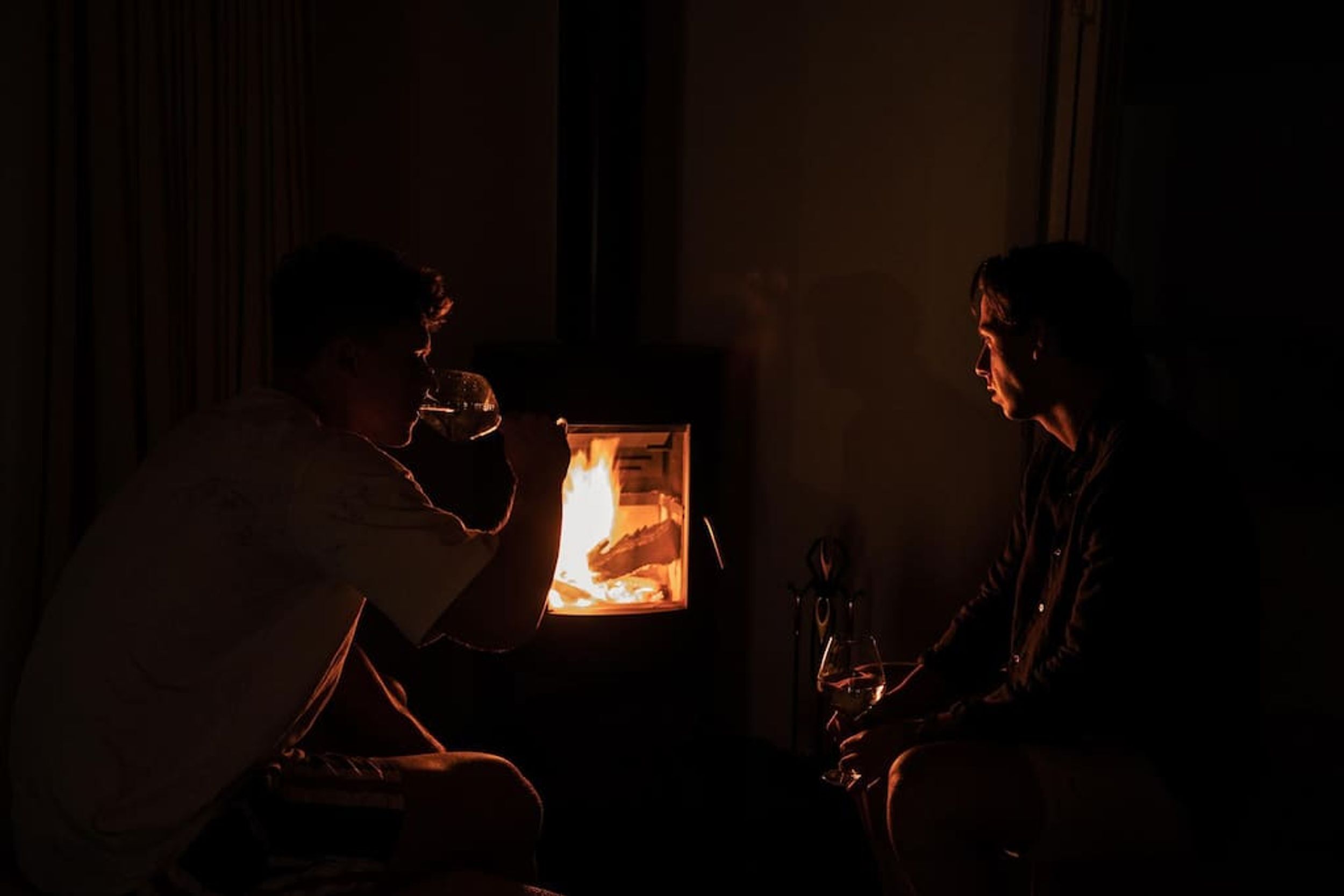 Two people sitting in a dimly lit room with a fireplace, one holding a glass.