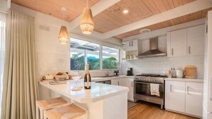 A modern kitchen with white cabinetry, a central island, bar stools, a large window, and a wooden ceiling. - Sol Coastal Retreat - Outdoor Spa and Sauna (Rosebud)