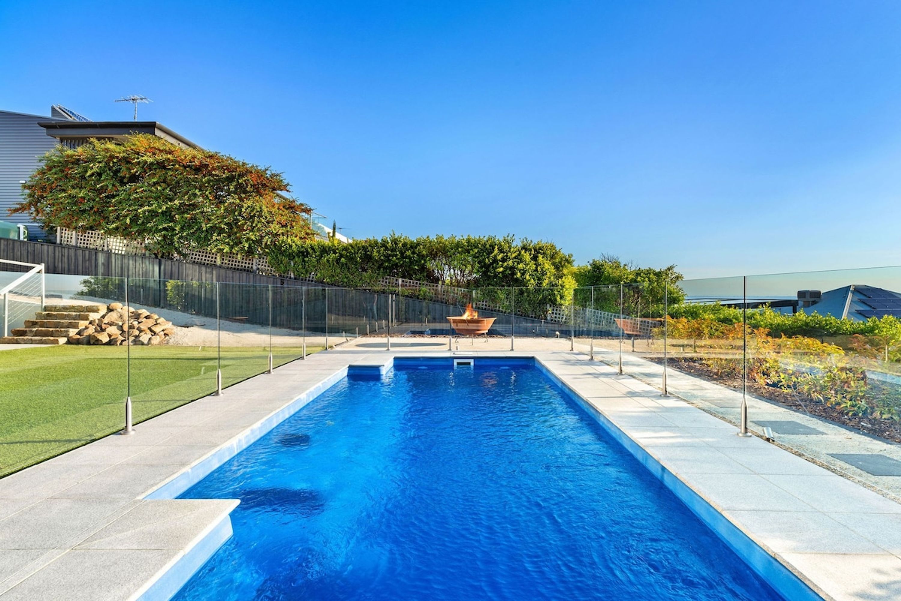 A rectangular swimming pool with a clear blue water surface, surrounded by a paved deck and a metal fence.