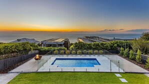 Una piscina con vista al océano al atardecer, rodeada de edificios modernos y un césped cuidado.
