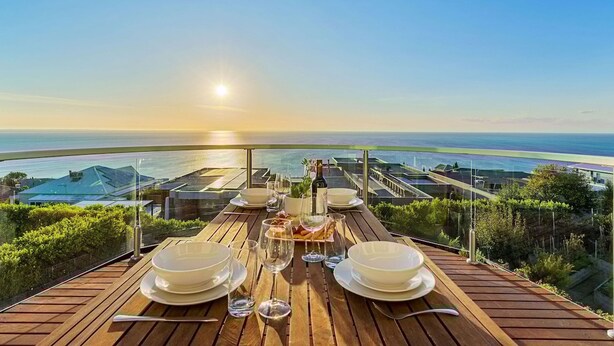 Una terraza de madera con una mesa dispuesta para una comida, con vista a una zona costera con edificios y cielo despejado.