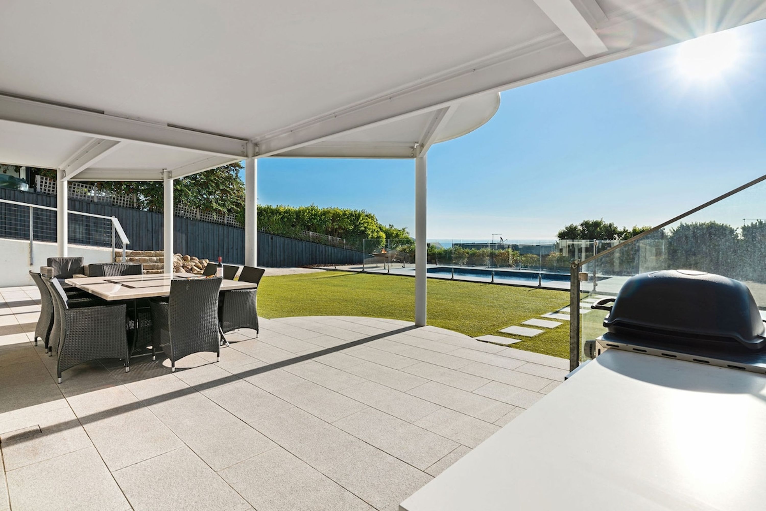 An outdoor patio with a dining table and chairs, a barbecue grill, and a view of a pool and greenery.
