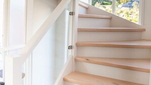 Una escalera con peldaños de madera y paredes blancas que conduce a una habitación luminosa con un amplio ventanal.