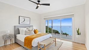 A bedroom with a bed, a ceiling fan, a large window with a view of the sea, and a side table with a lamp. - Clifftop Luxury With Spectacular Ocean Views (Mount Martha)