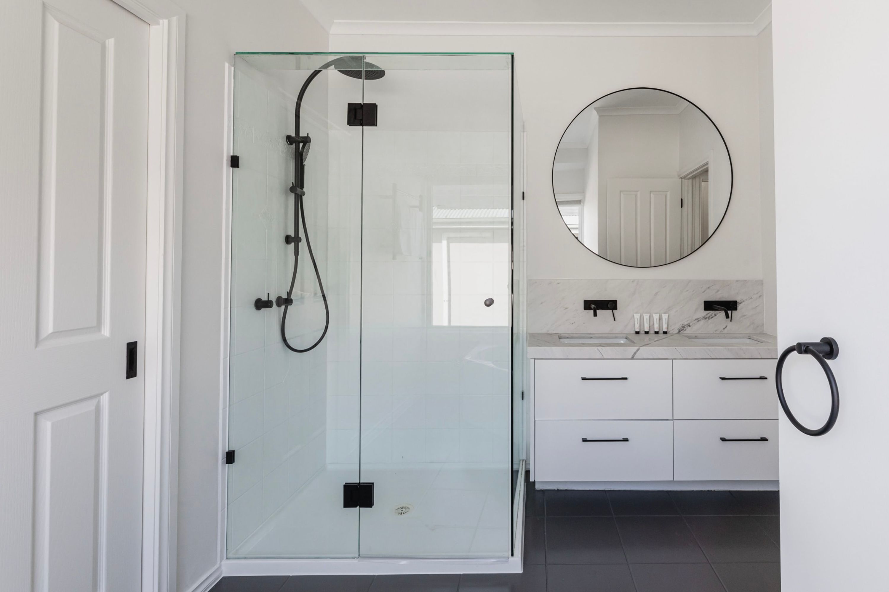 A modern bathroom with a glass-enclosed shower, a round mirror, and a white vanity with drawers and a sink.