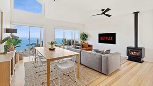 A spacious living room with a large window overlooking the sea, a fireplace, a ceiling fan, and a wall-mounted Netflix sign. - Clifftop Luxury With Spectacular Ocean Views (Mount Martha)