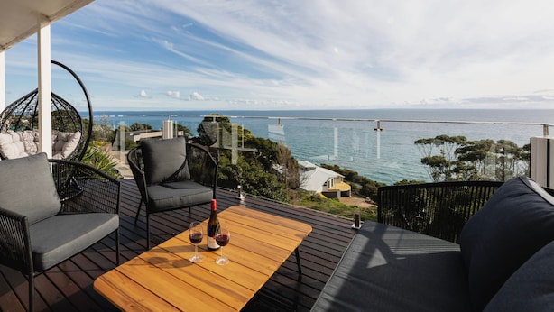 A balcony with a wooden table, two black chairs, and a hanging chair. The view includes a body of water and distant land. - Clifftop Luxury With Spectacular Ocean Views (Mount Martha)