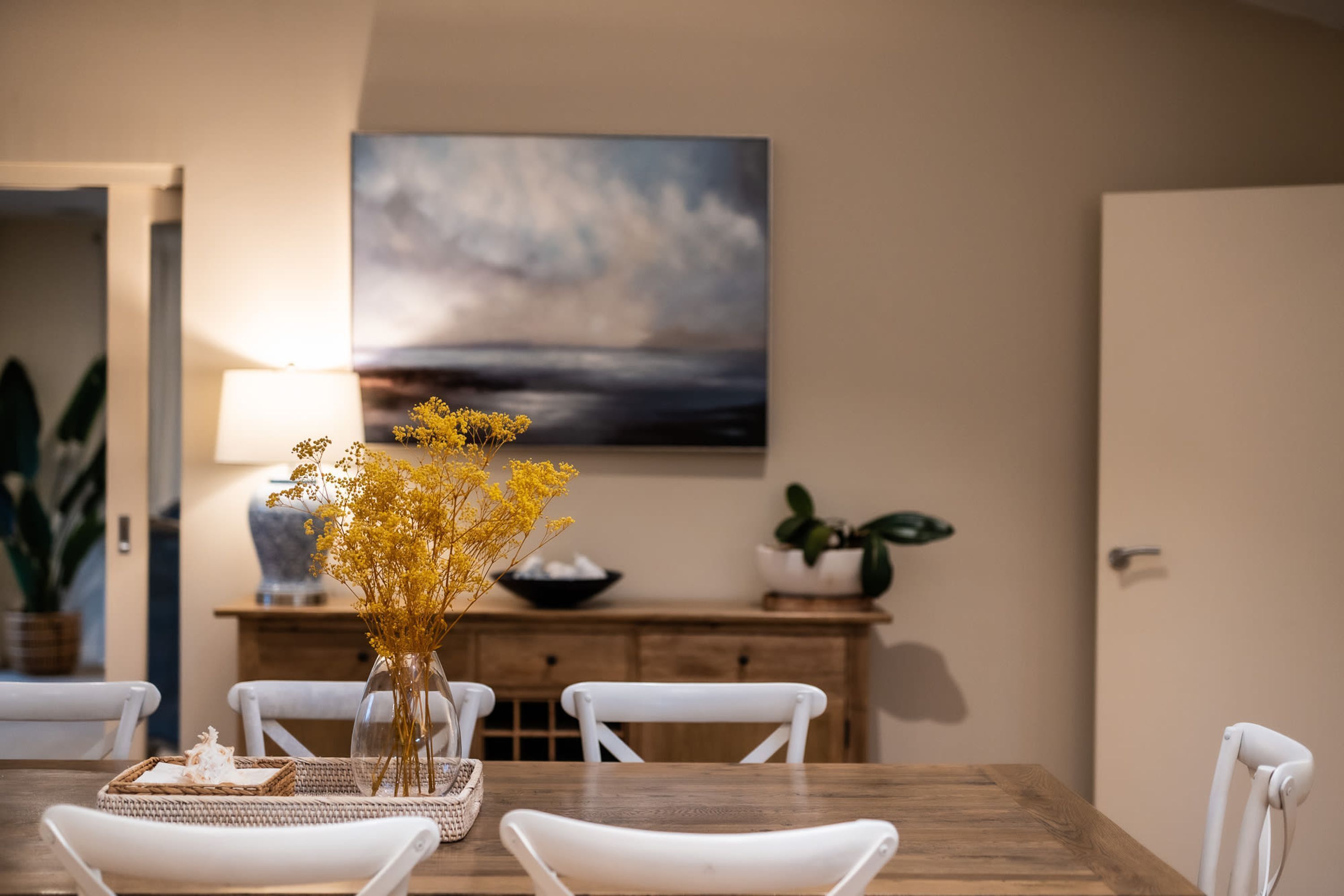 A dining area with a wooden table, white chairs, a vase with yellow flowers, a bowl, and a painting on the wall.