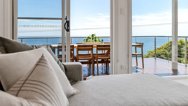 Un dormitorio con una cama grande, vista al mar a través de una puerta corrediza de vidrio y una terraza de madera con una mesa y sillas.