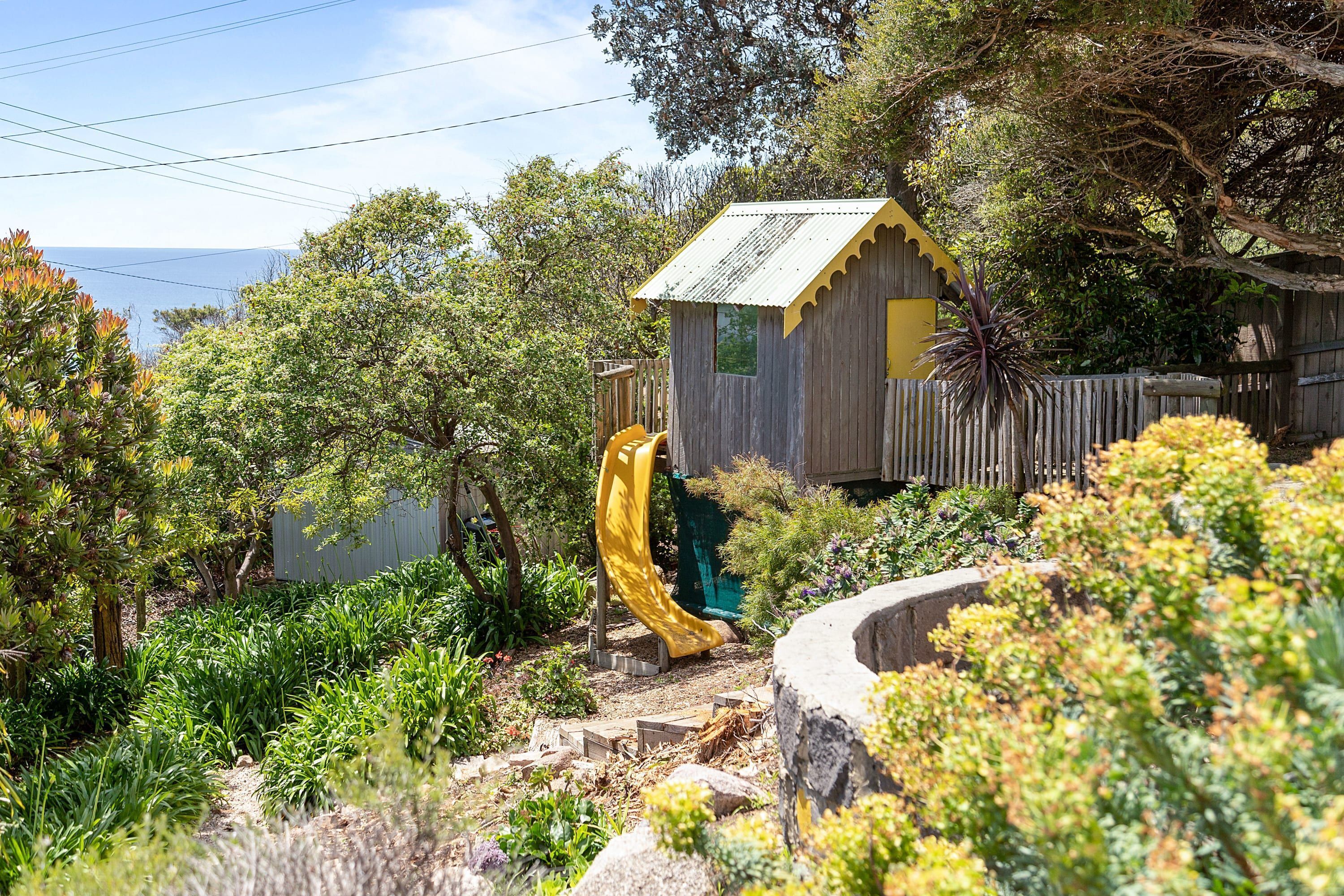 A wooden playhouse with a yellow slide, surrounded by lush greenery and trees.