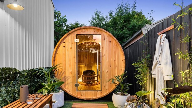 Une structure de sauna en bois, dotée d’une porte, d’un banc en bois, de plantes en pot et d’une serviette blanche accrochée à une clôture en bois.