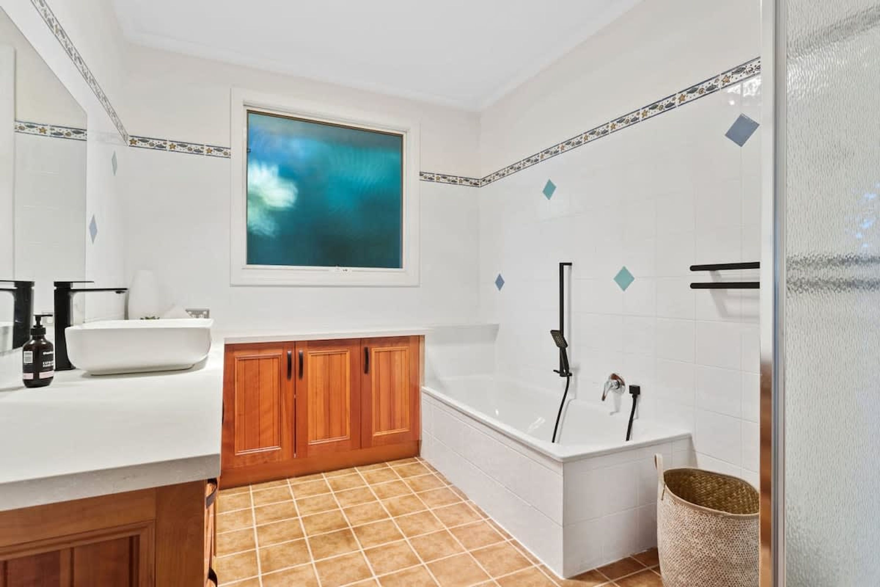 A modern bathroom with a white bathtub, wooden cabinets, a large window, and a tiled floor.