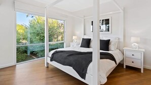 A modern bedroom with a four-poster bed, a large window with a view of greenery, and a bedside table with a lamp. - Blairgowrie Beachside Retreat Solar Heated Pool (Blairgowrie)