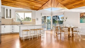 A modern kitchen with wooden flooring, a central island with stools, a dining area with a table and chairs, and a large window providing natural light. - Blairgowrie Beachside Retreat Solar Heated Pool (Blairgowrie)