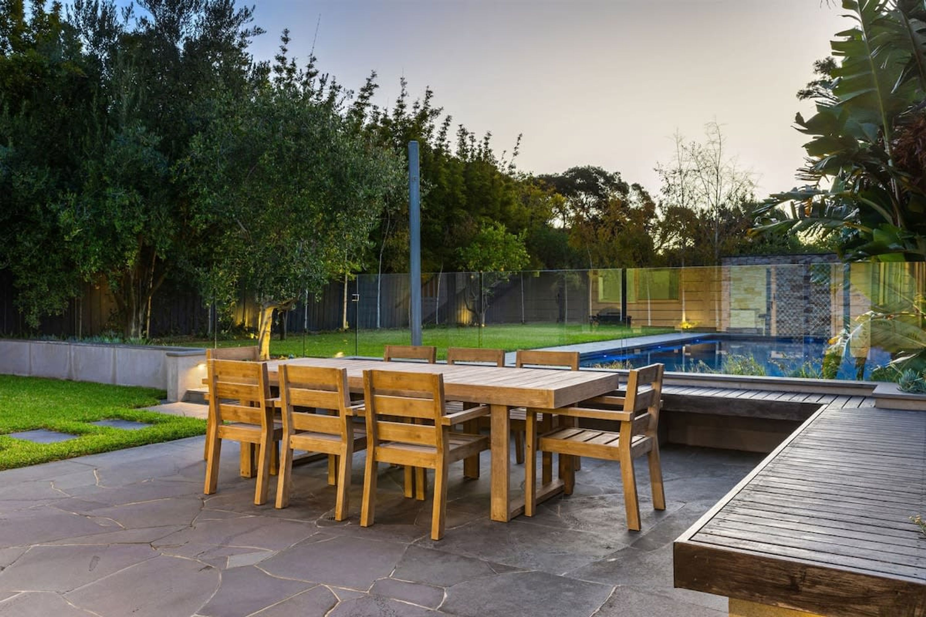An outdoor patio with a wooden table and chairs, a stone floor, and a swimming pool in the background.