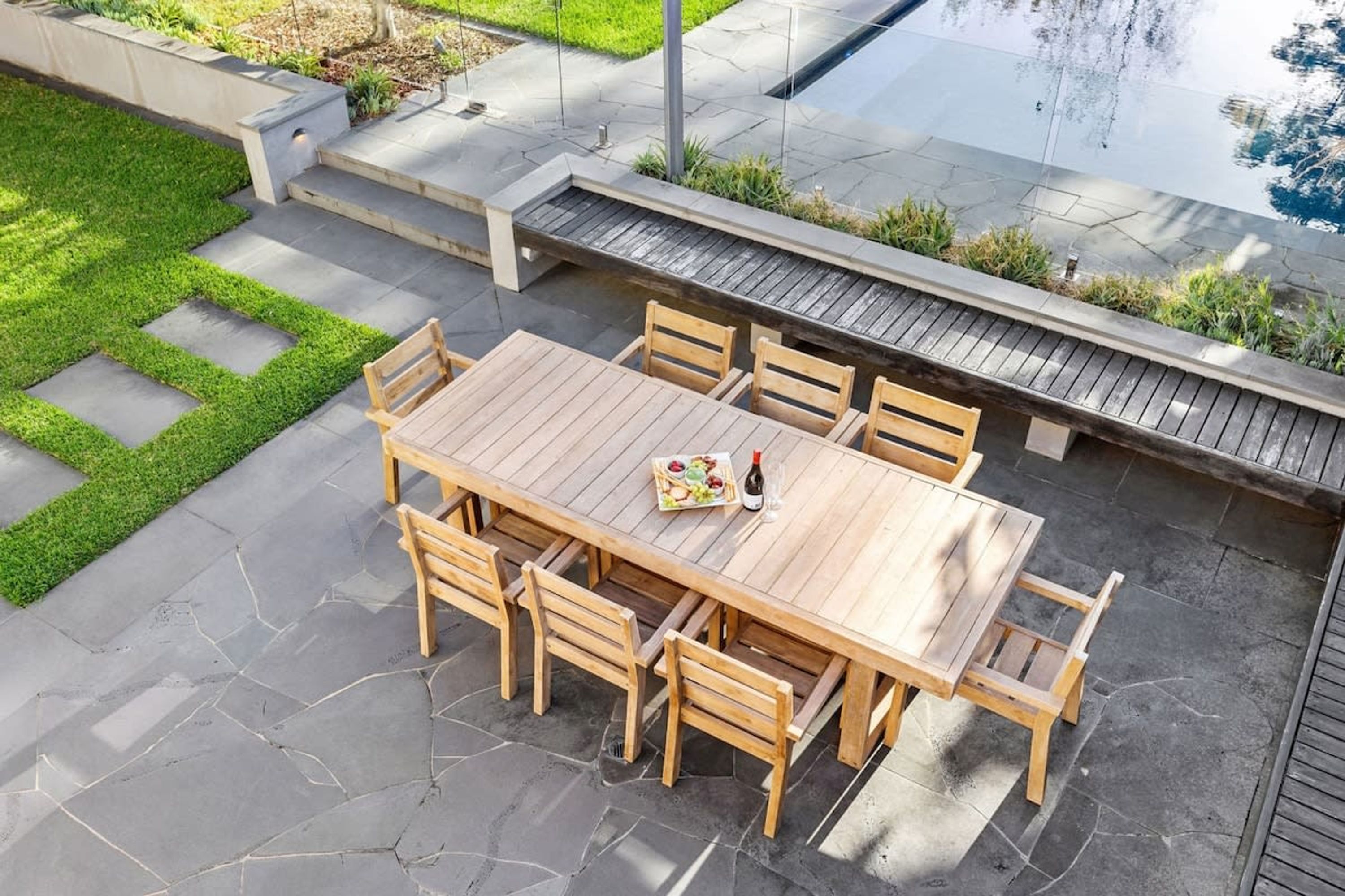 A wooden outdoor dining area with a table and chairs, surrounded by a stone patio and a small grassy area.